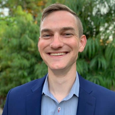 Smiling headshot of Nate, wearing a blue suit jacket, in front of some shrubberies