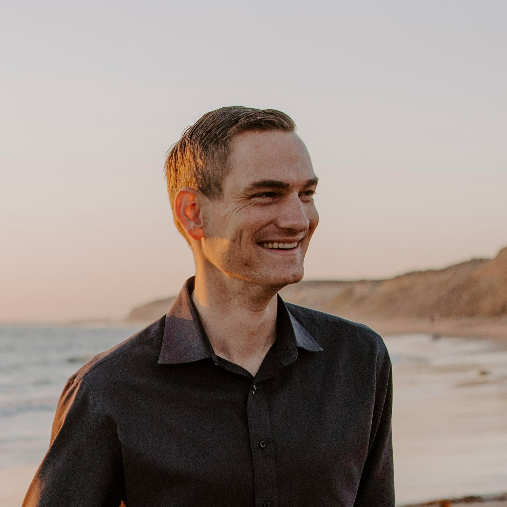 A portrait of Nate smiling, looking off to his left, with the ocean in the background.